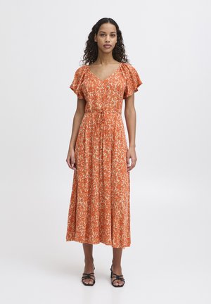 Kobieta stoi, ubrana w pomarańczową, kwiecistą sukienkę midi z krótkimi rękawami, dekoltem w serek i wiązaniem w talii, w połączeniu z czarnymi sandałami na paski.