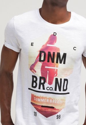 Homme portant un T-shirt blanc avec un graphique d'une femme tenant une planche de surf au coucher de soleil et le texte "DNM Brand Co. Summer Breeze 1998."