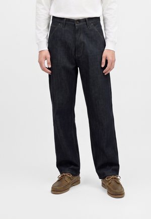 Personne portant un jean droit foncé, une chemise blanche à manches longues et des mocassins marron à lacets sur un fond uni.