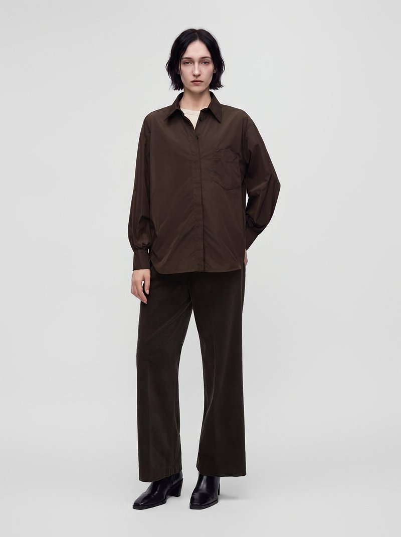 Femme aux cheveux courts et foncés portant une chemise marron foncé boutonnée, un pantalon large assorti et des bottines noires sur un fond clair uni.