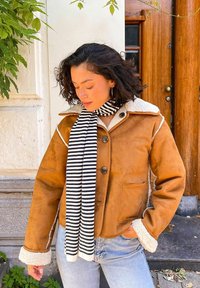 Bruine suède jas met witte schapenvacht kraag en manchetten, met een knoopsluiting en twee voorzakken. Draag met een zwart-wit gestreepte sjaal.