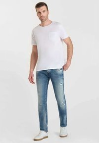 Witte t-shirt met korte mouwen en een zakje, gecombineerd met lichtblauwe distressed jeans en witte sneakers, staand tegen een neutrale achtergrond.
