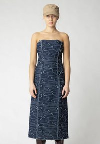 Jeans trägerloses Kleid mit wellenförmigen hellblauen Mustern, ausgefransten Kanten und gerader Silhouette. Kombiniert mit einer beigen Kappe.