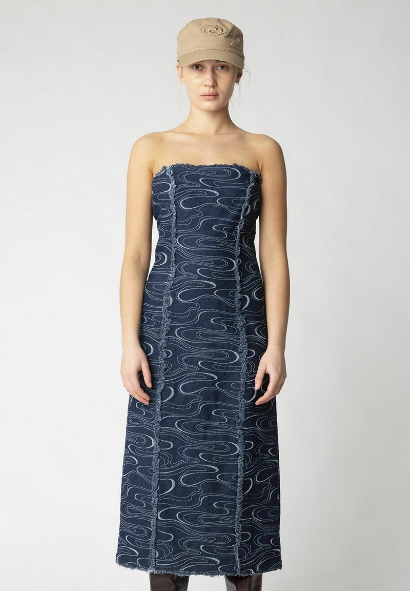 Jeans trägerloses Kleid mit wellenförmigen hellblauen Mustern, ausgefransten Kanten und gerader Silhouette. Kombiniert mit einer beigen Kappe.