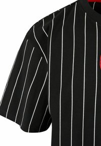 Svart bomullströja med vita vertikala ränder, med rund halsringning och en liten röd emblem på framsidan. Korta ärmar.