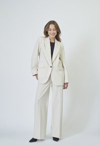 Cream-colored, structured blazer with two front pockets and a single button, paired with wide-leg cream pants and black heels.