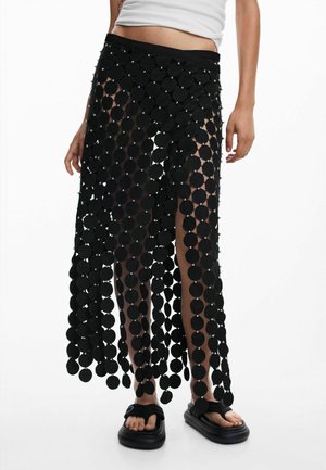 Persoană îmbrăcată cu o fustă neagră formată din discuri circulare mari interconectate peste pantaloni scurți negri, asortată cu sandale negre și un top alb scurt.