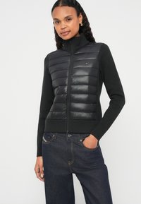 Μαύρο μπουφάν τύπου puffer με φερμουάρ μπροστά, διαθέτοντας πλεκτή ριπ υφή στο κάτω μέρος, μακριά μανίκια και αντίθετη υφή μεταξύ των υλικών.