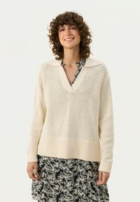 Maglione lavorato a maglia color crema con scollatura a V, caratterizzato da una trama aperta e un orlo a coste. Indossato sopra un vestito nero a fiori.