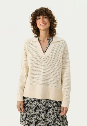 Cremefarvet strikket sweater med V-udskæring, der har en tekstureret åben vævning og en ribbet kant. Båret over en blomsterprintet sort kjole.