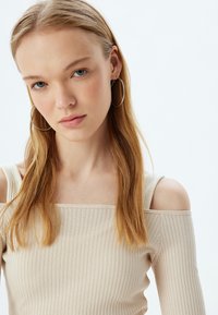 Top beige a costine off-shoulder con scollatura quadrata e spalline sottili, caratterizzato da una texture morbida. La modella indossa grandi orecchini a cerchio in argento.