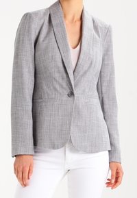 Šedý strukturovaný blazer s elegantním střihem, zapínáním na jeden knoflík a klopami. Nosený přes světlejší top a kombinovaný s bílými kalhotami.