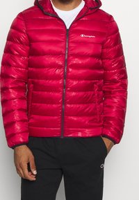 Rød puffjakke med horisontalt quiltet design, høj krave, lynlås lukning og Champion-logo på venstre bryst. Fleece-fodrede manchetter.