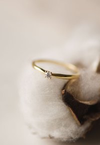 Gouden ring met een slanke band en een kleine, ronde, heldere edelsteen die in een vierpuntszetting is gezet, rustend op zachte katoen.