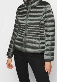 Polstrad grön puffjacka med hög krage, dragkedja fram och ribbade ärmar. Utmärkande horisontell sömnad och glänsande textur.