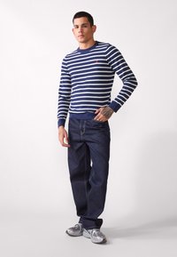 Marineblauwe en witte gestreepte gebreide trui met een ronde hals; gedragen met donkerdenim jeans en grijze sneakers. Heeft ribgebreide manchetten en een zoom.