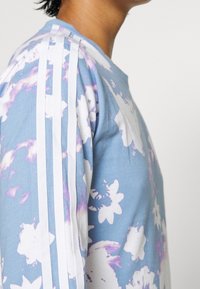 Ljusblå långärmad skjorta med vitt blommönster, med tre vita ränder längs axlarna. Mjuk bomullstextur.