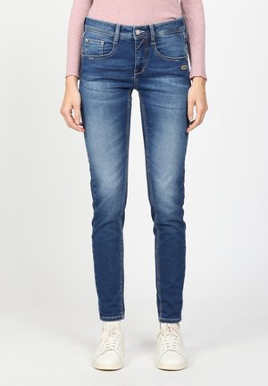 Dünne blaue Jeans mit einem verblassten Waschung, mit fünf Taschen, einem Knopfverschluss und kontrastierenden Nähten. Getragen mit weißen Turnschuhen.