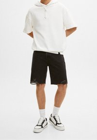 Sudadera blanca con capucha, pantalones cortos de mezclilla negros con dobladillo deshilachado y zapatillas en blanco y negro. El atuendo presenta materiales casuales y cómodos.