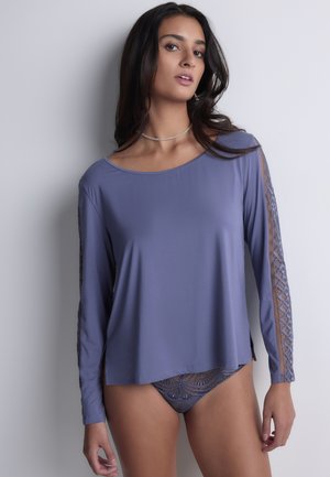 Femme portant un haut manches longues lavande avec des bandes en dentelle sur les manches et une lingerie en dentelle assortie, debout devant un fond neutre.