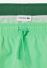 Grüne Badebermuda mit einem Kordelzug, elastischem Bund und einem Lacoste-Logo-Tag. Glatte Textur mit leichtem Design.