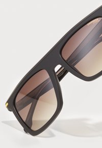 Ochelari de soare rectangulari negri cu lentile degrade de culoare marron, finisaj mat și accente subtile de aur pe ramă. Design elegant, unghiular.