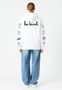 Bela jopa s kapuco z napisom "bodi prijazen." v črni na hrbtni strani. Ima barvite grafike in besedilo na rokavih. Kombinirana z modrimi širokimi kavbojkami.