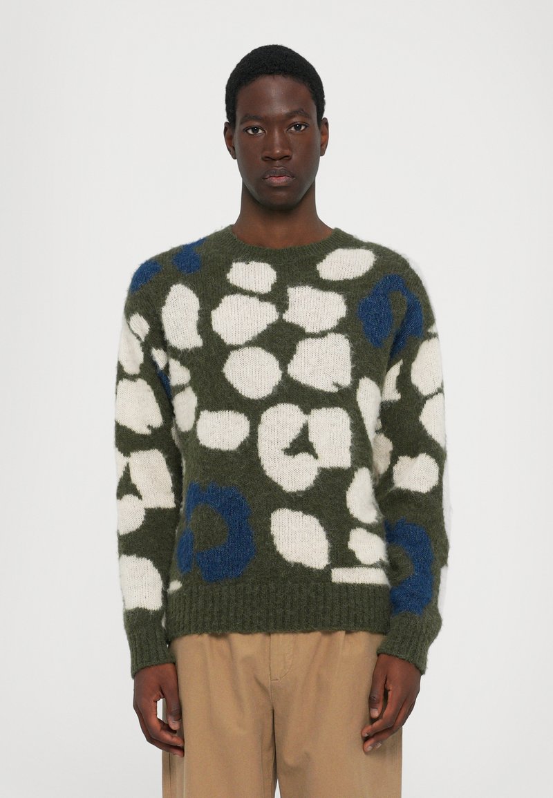 Suéter verde con patrones abstractos en blanco y azul, tejido texturizado, cuello redondo, puños y dobladillo acanalados, llevado sobre pantalones color beige.