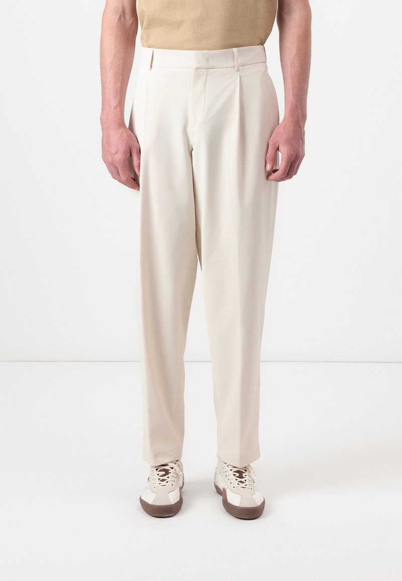Beige bukser med skræddersyet pasform, med høj talje, frontplisseringer og en glat stofstruktur. Båret med hvide sneakers.