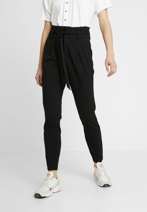 Person iført sorte højtaljede bukser med tapering, papirlommer i taljen og bundet bælte, kombineret med hvide sneakers og en kortærmet hvid skjorte med knapper.