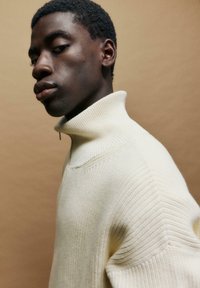 Cremfarbener Rollkragenpullover mit geripptem Muster, einem Reißverschlusskragen und lockerem Schnitt; zeigt subtile Strickmuster im Stoff.