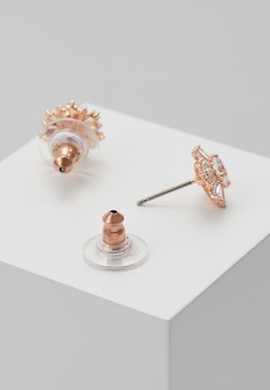 Boucles d’oreilles clous en or rose avec plastique transparent et attaches en or rose présentées sur une surface blanche.