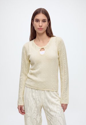 Jeune femme aux cheveux bruns et raides portant un haut en maille crème à manches longues avec un détail en forme de trou et un pantalon assorti à motifs, sur un fond uni.