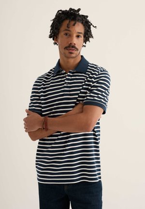 Göndör hajú férfi, sötétkék és fehér csíkos galléros pólót és sötét farmert visel, karját keresztbe téve áll sima háttér előtt.