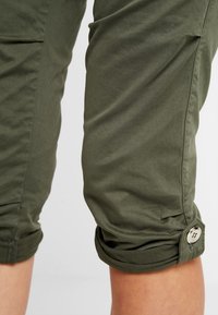 Olivově zelené capri kalhoty vyrobené z lehké tkaniny, s rolled hem a knoflíkovými detaily po stranách. Hladká textura a ležérní design.