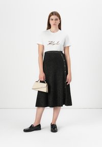 Modelo con una camiseta blanca que lleva el texto "Karl", combinada con una falda negra brillante y sosteniendo un bolso beige, contra un fondo neutro.