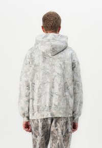 Un uomo di spalle indossa una felpa con cappuccio e pantaloni con un abbinamento di modelli mimetici grigio chiaro e marrone, raffiguranti rami e foglie.