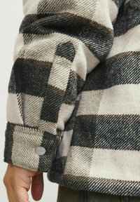 Giacca di flanella con un motivo a quadri nelle tonalità di beige, nero e oliva. Include polsini cuciti e un dettaglio con bottone in metallo.