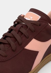 Maroon suede sneaker featuring brown laces, a pink accent logo, and a light pink panel. Textured surface with a gum rubber sole.