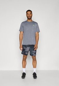 Grå atletisk skjorte med teksturert mønster og grønn logo, kombinert med mørke tie-dye shorts. Modell iført svarte joggesko og hvite sokker.