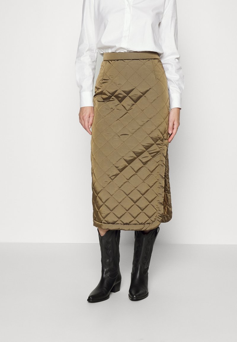 Frau in einem olivgrünen, gesteppte Midirock mit einem seitlichen Schlitz, weißem Langarmshirt und schwarzen Lederstiefeln bis zur Mitte der Wade.