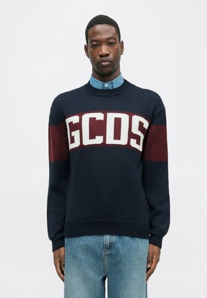 Jūras spēku adīts džemperis ar bordo un baltām horizontālām svītrām, kas rotāts ar "GCDS" logo. Ar apaļu kakla izgriezumu un ribbētiem piedurknes galiem, kombinēts ar gaiši zilu krekla.