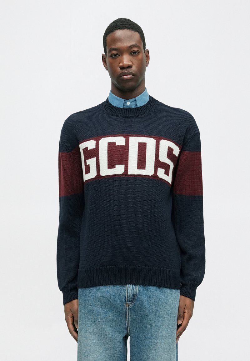 Tumesinine kootud sviiter marooni ja valgete horisontaalsete triipudega, millel on "GCDS" logo. Ümmargune kaelus ja soonikkoes varrukad, koos helesinise särgiga.