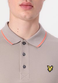 Světle šedá polo tričko s texturovanou tkaninou, s oranžovým lemem na límci, třemi tmavými knoflíky a žlutým vyšitým logem.