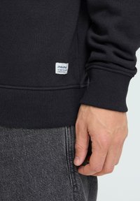 Sweat-shirt noir en coton avec poignets et ourlet côtelés. Présente une petite étiquette rectangulaire sur le côté avec un texte blanc. Pantalon gris visible en dessous.