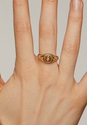 Anillo de oro con forma ovalada, con un centro elevado y pequeños acentos de diamante a cada lado, colocado sobre una mano con un tono de piel claro.