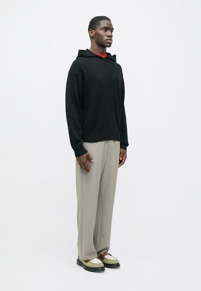 Bărbat stând de profilen, îmbrăcat într-o bluză cu glugă neagră, pantaloni bej largi și pantofi loafers bej-maro pe un fundal alb simplu.