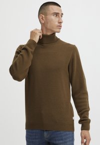 Pull col roulé marron en tissu doux, avec des poignets et un ourlet côtelés. Porté avec un jean bleu, mettant en valeur une coupe ajustée.