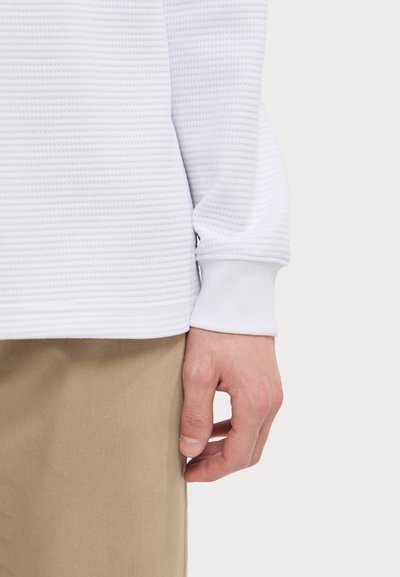 White textured long-sleeve shirt with ribbed cuffs, paired with beige pants; focus on shirt's fabric and fit along the wrist and waist.