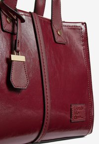 Burgundi odinė rankinė su lygiu paviršiumi, auksinės spalvos detalėmis ir siuvinėjimo akcentais. Šone yra logotipo ženklas.